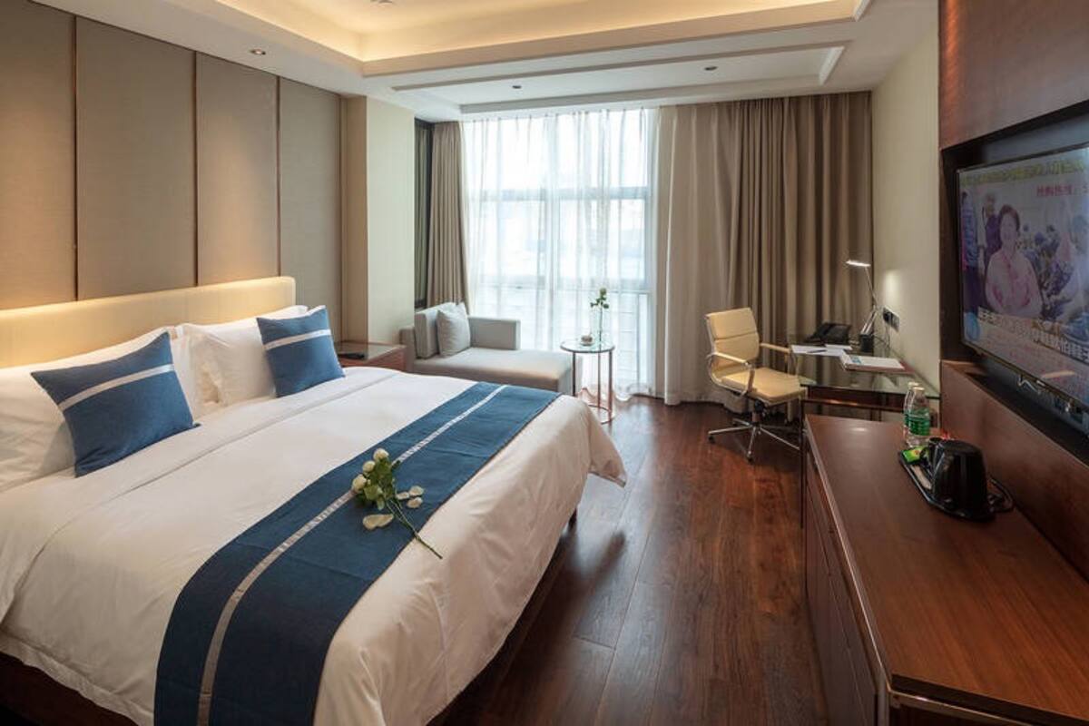 Habitación de hotel con una cama grande, un escritorio, una silla, un televisor y una pequeña mesa con una flor.