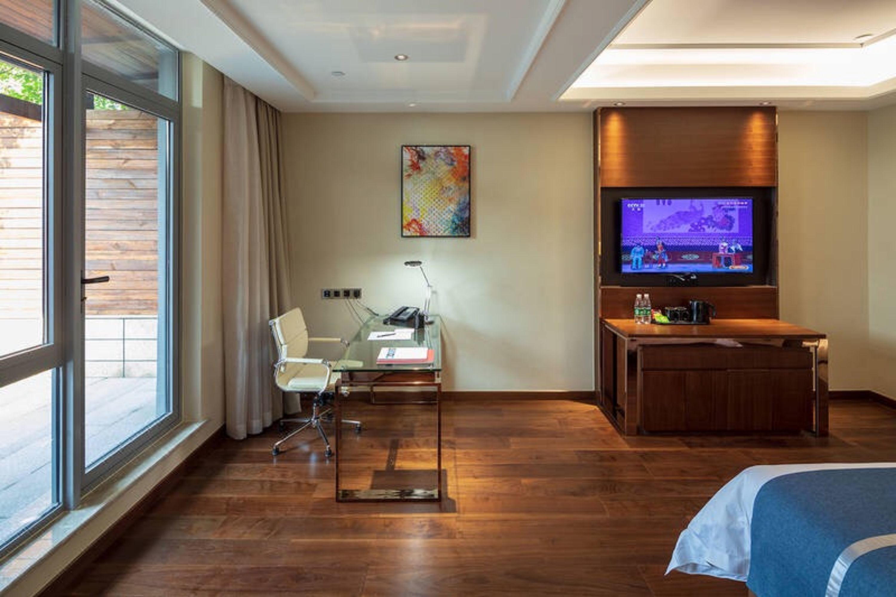 A hotel room with a large window, a desk with a chair, a television, and a bed.
