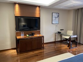 Habitación de hotel moderna con un televisor de pantalla plana montado en un panel de madera, una mesa de centro de vidrio con un control remoto, una silla blanca y un pequeño escritorio con una lámpara.