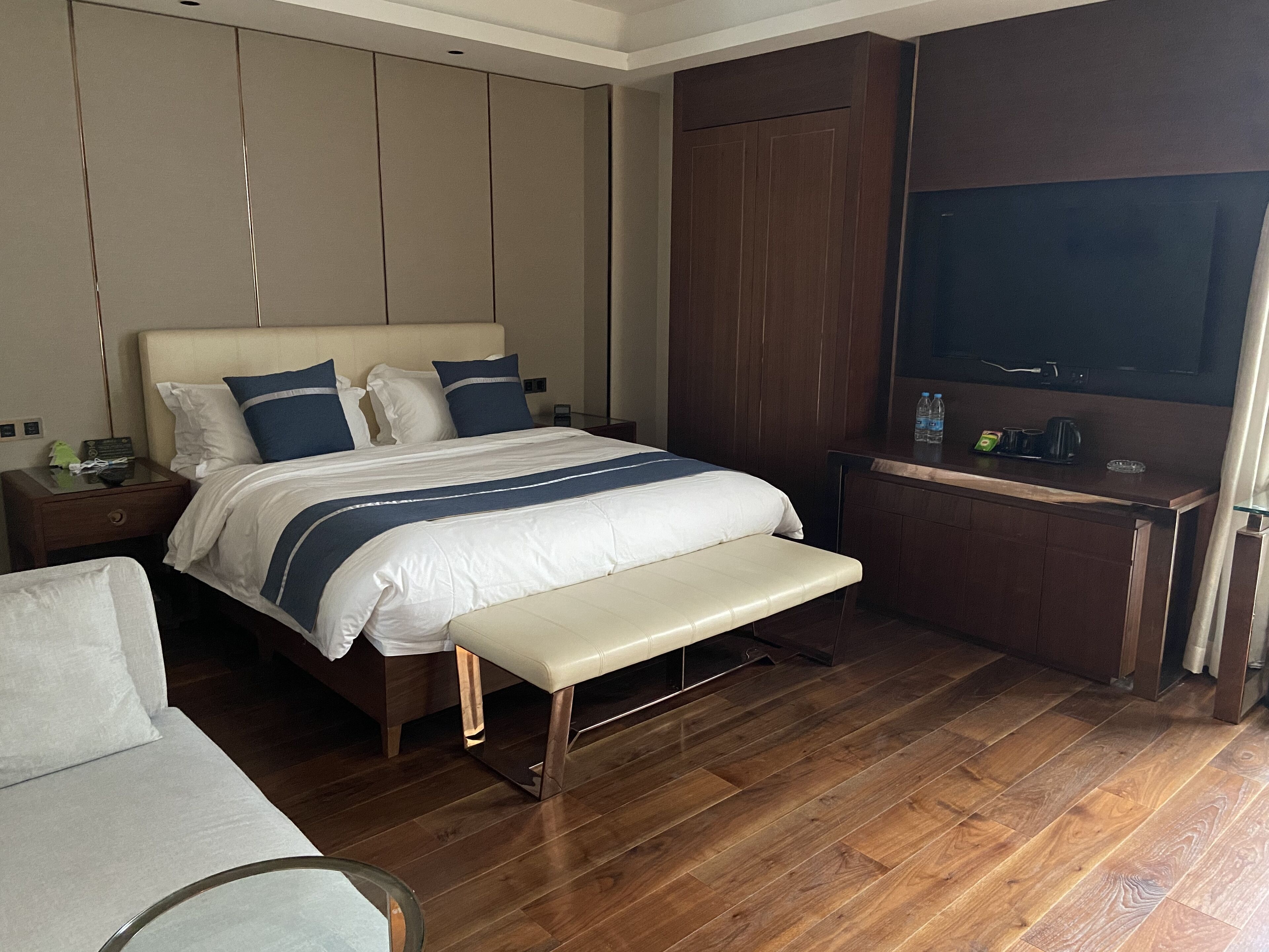 A modern hotel room with a large bed, wooden flooring, a bench, a desk with a flat-screen TV, and a small table with a water bottle and a cup.