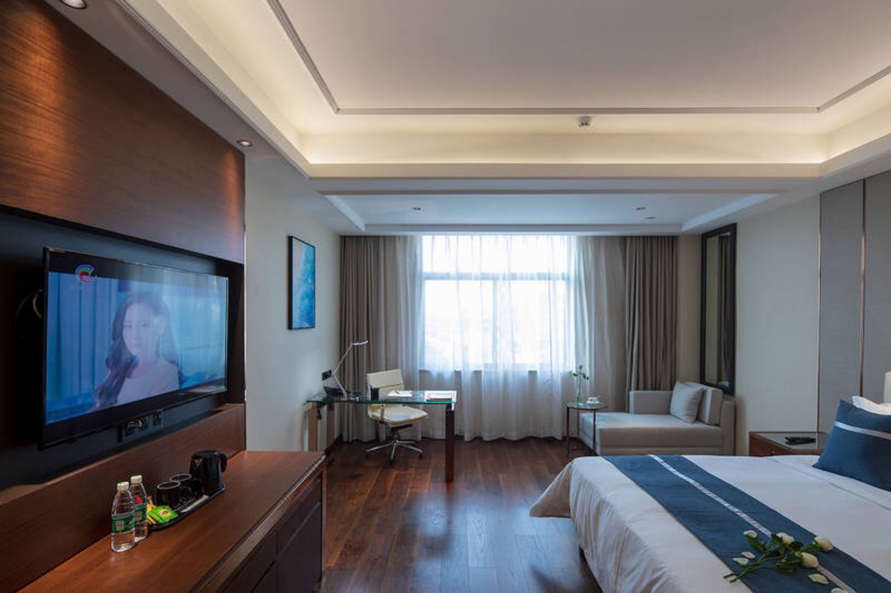 A hotel room with a large flat-screen TV, a desk with a chair, a bed, a sofa, and a window with curtains.