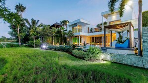 A modern two-story house with a glass balcony, surrounded by lush greenery and palm trees. - Phocea Golf View Villa by Premier Hospitality Asia (Pecatu)