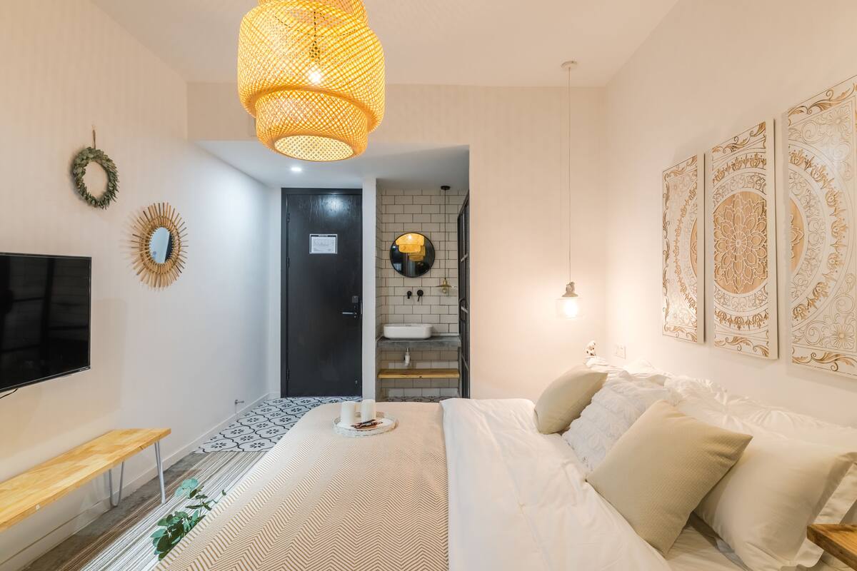 Ein modernes Schlafzimmer mit einem großen Bett, einem Fernseher, einer hölzernen Sitzbank, einem dekorativen Spiegel und einem einzigartigen Wandkunstwerk.