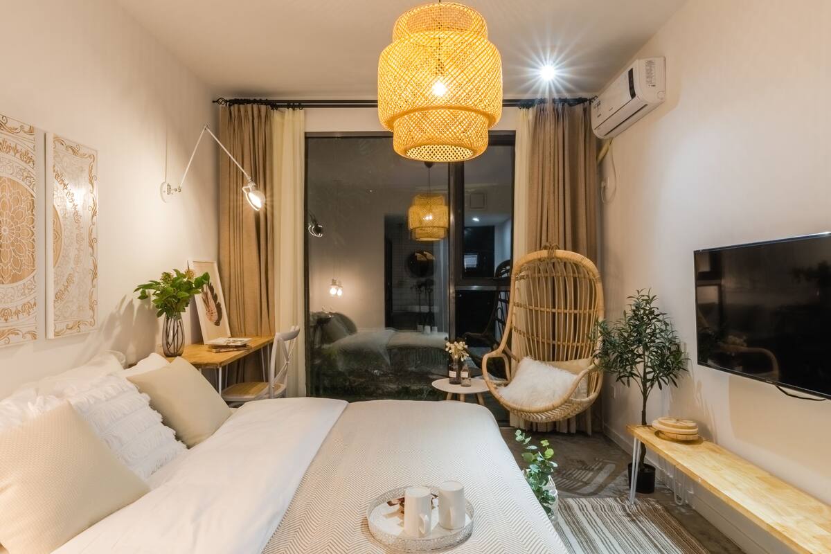 Ein Hotelzimmer mit Bett, Fernseher, Korbsessel, kleinem Tisch mit Getränken und großem Fenster mit Vorhängen.