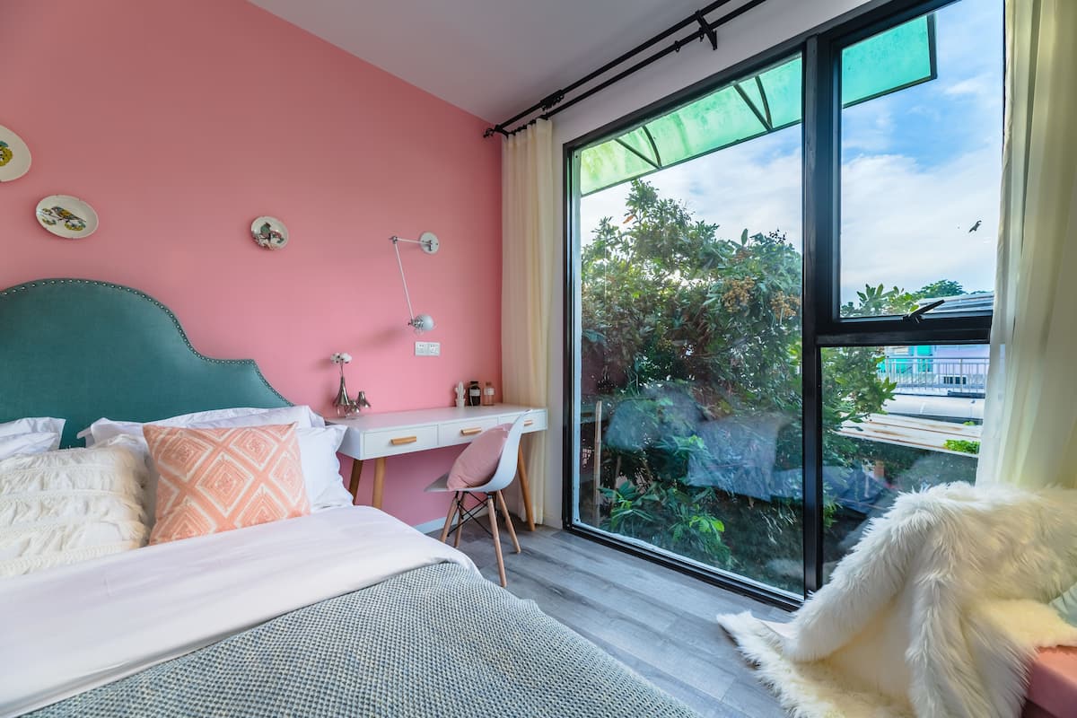 Ein Schlafzimmer mit einem Bett, einem Schreibtisch und einem Stuhl. Ein Fenster bietet Blick auf grüne Vegetation.