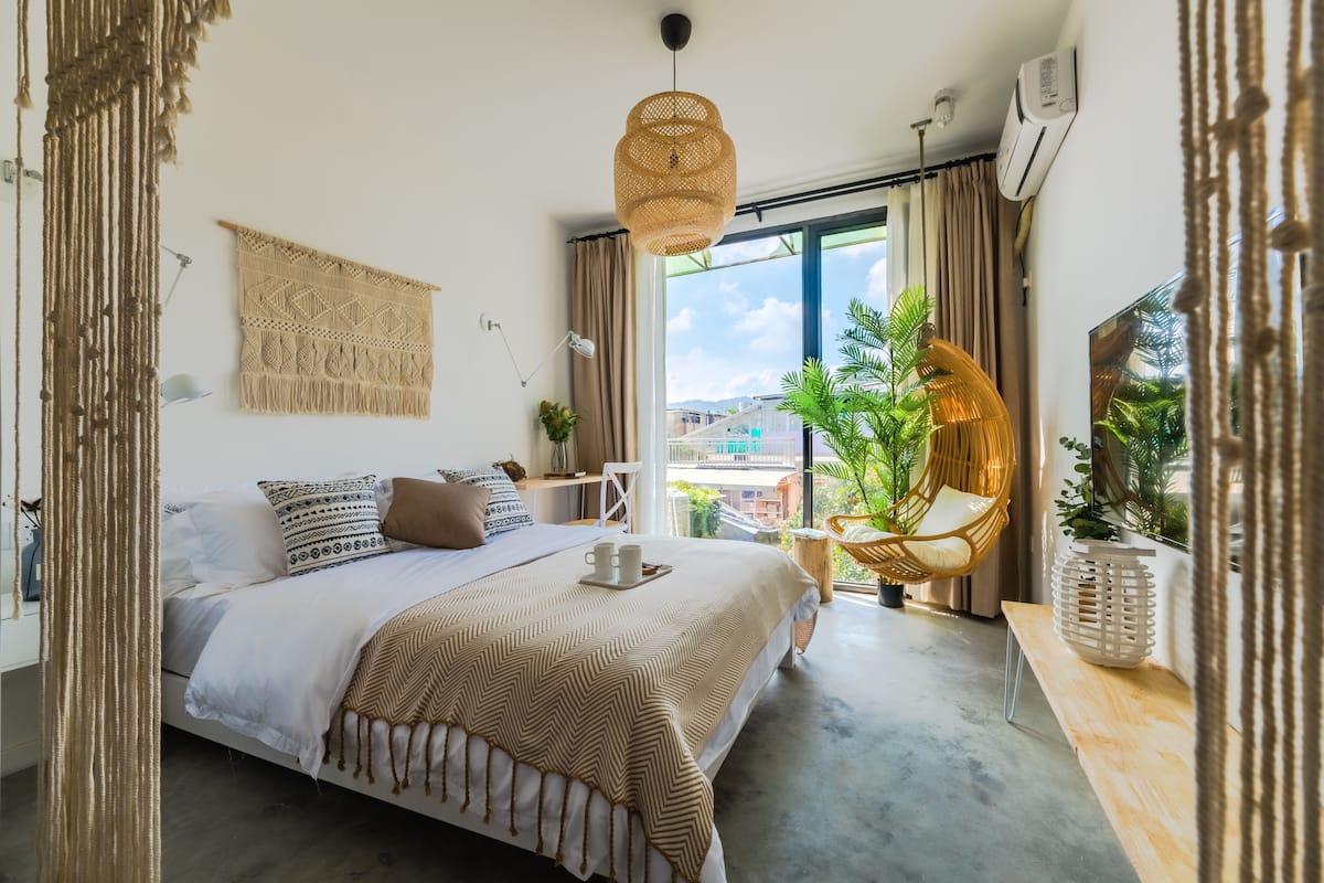 Ein modernes Schlafzimmer mit Bett, Nachttisch, hängender Makramee-Dekoration und Blick auf Grünfläche.