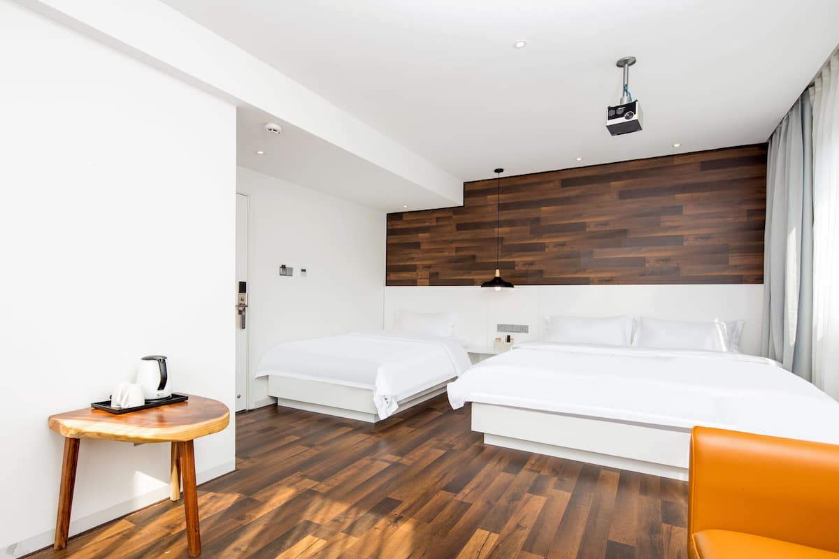 A modern hotel room with two beds, a wooden floor, a small table with a kettle, and a bright overhead light.