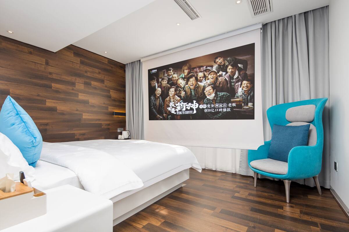 A modern hotel room with a large bed, a blue armchair, a wooden wall panel, and a movie poster on the wall.