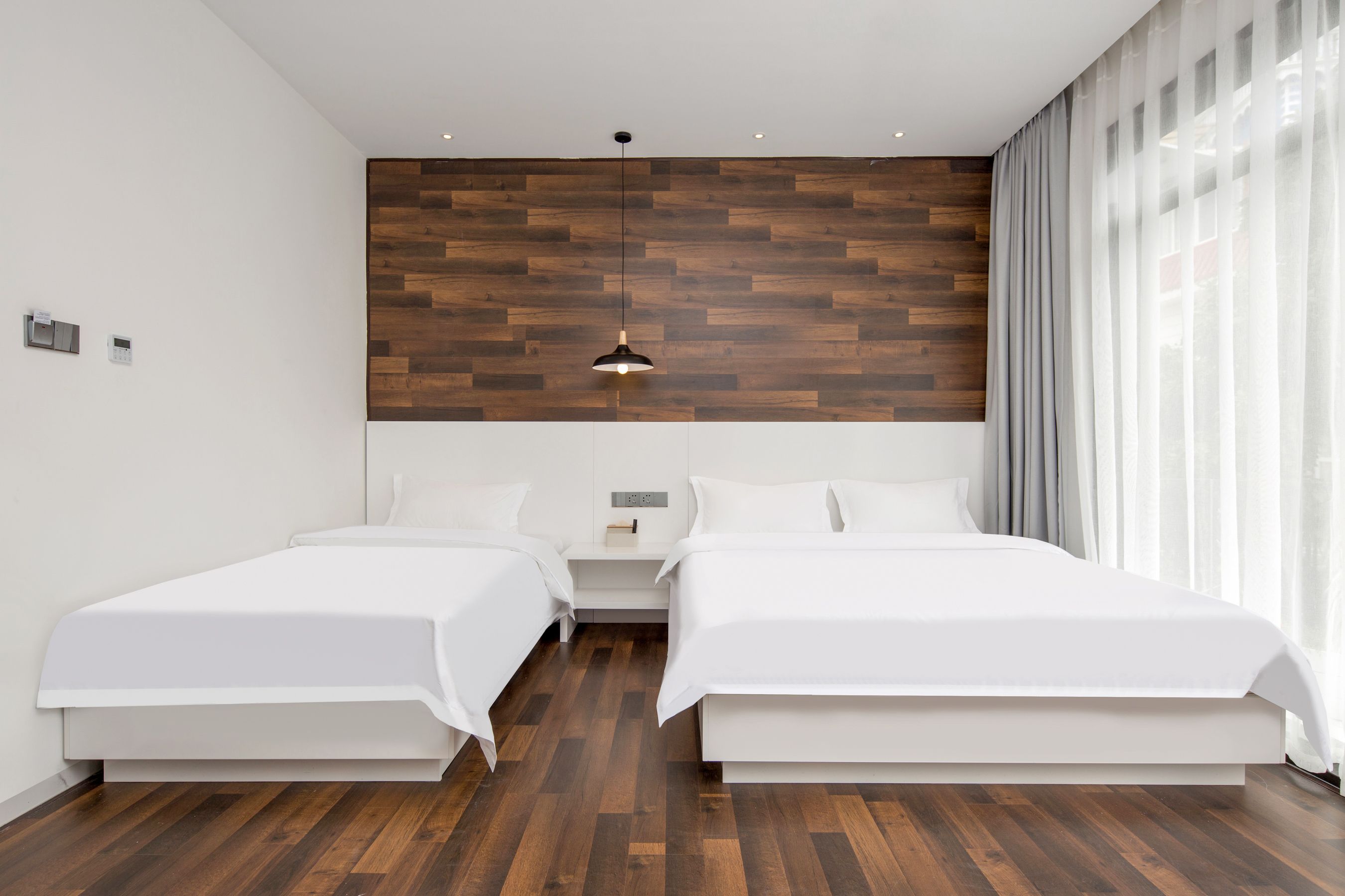 A modern hotel room with two beds, a wooden accent wall, a hanging pendant light, and a large window with curtains.