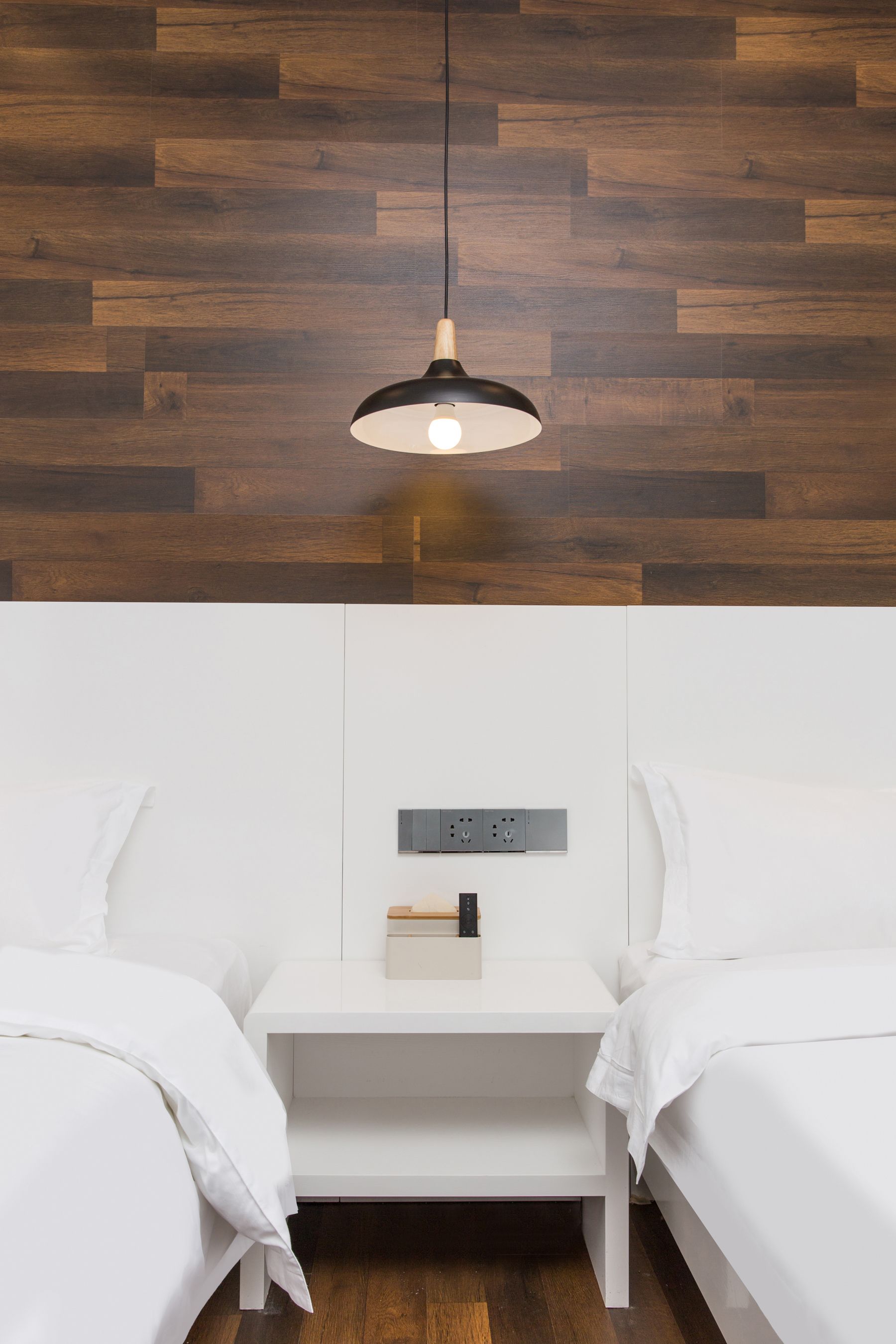 A modern bedroom with a wooden floor, white bedding, a bedside table with a lamp, and a power outlet.