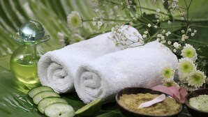 A spa setting with a rolled white towel, cucumber slices, a glass bottle, and bowls of bath salts on a green leaf.