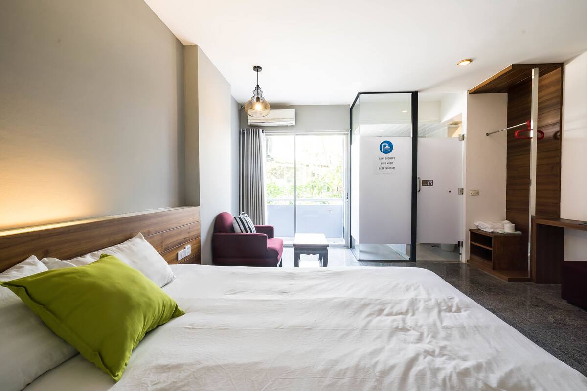 Une chambre d’hôtel avec un grand lit, un fauteuil rouge, une petite table et une porte coulissante donnant sur un balcon.
