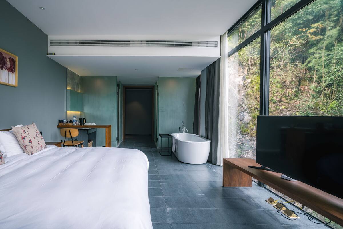 A modern hotel room with a large bed, a desk, a bathtub, and a TV.