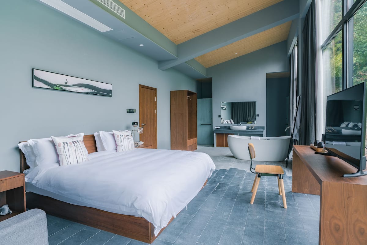 A modern bedroom with a large bed, a wooden chair, a bathtub, and a TV.