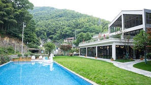 A modern building with a swimming pool, surrounded by a well-maintained lawn and a forested hillside.