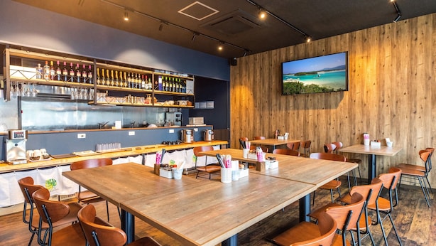 Un restaurante con mesas y sillas de madera, una zona de bar con botellas y vasos, y una gran pantalla de televisión en la pared.