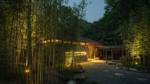 Un bâtiment moderne à la façade en pierre, entouré de bambous et bordé d’un chemin de gravier menant à l’entrée.