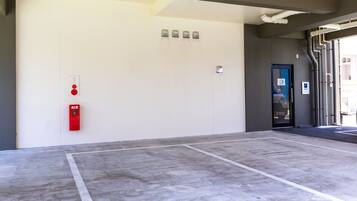 Un hall d’entrée propre et moderne, avec un détecteur d’incendie et un panneau indiquant la sortie de secours sur le mur, un sol en béton avec des marquages au sol pour le stationnement, et une porte donnant accès à un autre espace.