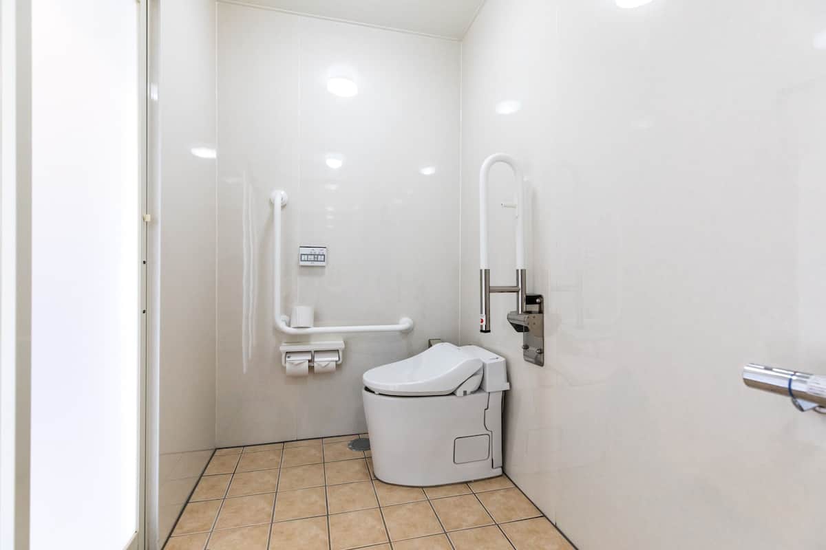 Un baño pequeño y blanco, con azulejos, un inodoro, un pasamanos y un espejo instalado en la pared.
