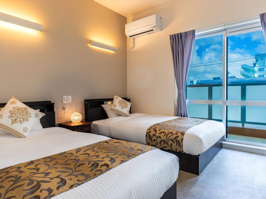 A hotel room with two beds, a nightstand with a lamp, an air conditioning unit, and a large window with curtains.