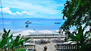 Eine Terrasse direkt am Strand mit Steinboden, Liegestühlen und Sonnenschirmen mit Blick auf das Meer und Boote.