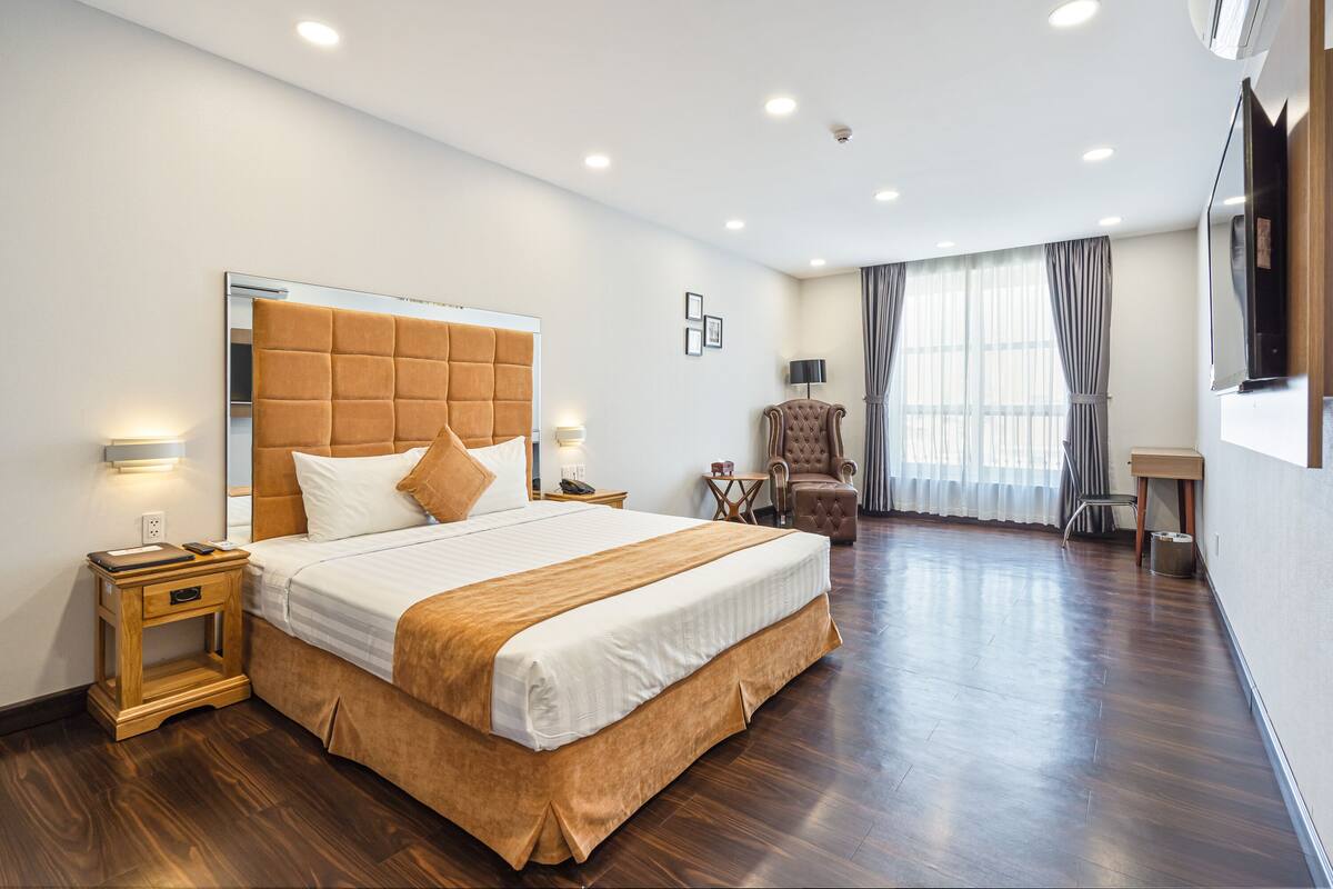 Quarto de hotel com uma cama grande, mesas de cabeceira de madeira, uma cadeira, uma escrivaninha e uma TV fixada na parede.