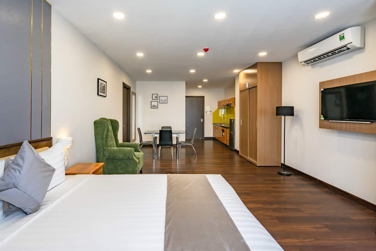 Quarto de hotel com cama, uma poltrona verde, uma mesa de jantar com cadeiras, uma cozinha e uma TV fixada na parede.