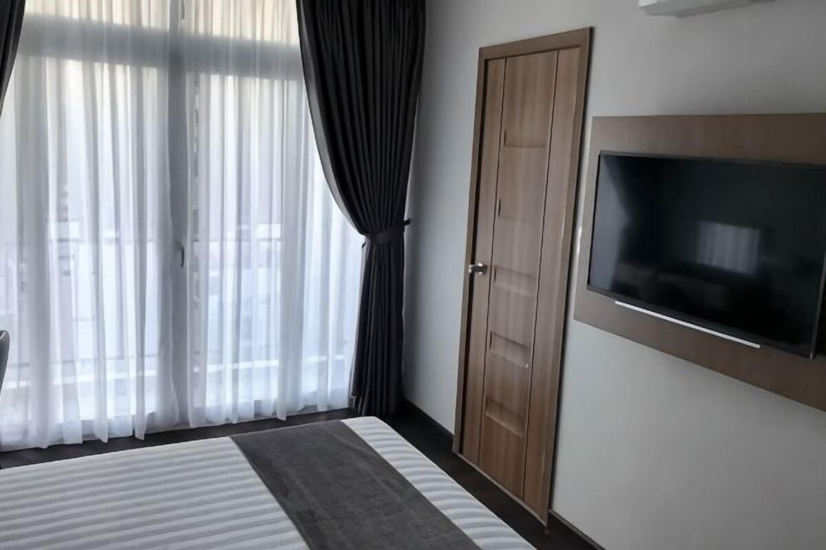 A hotel room with a bed, a TV mounted on the wall, a wooden door, and sheer curtains.