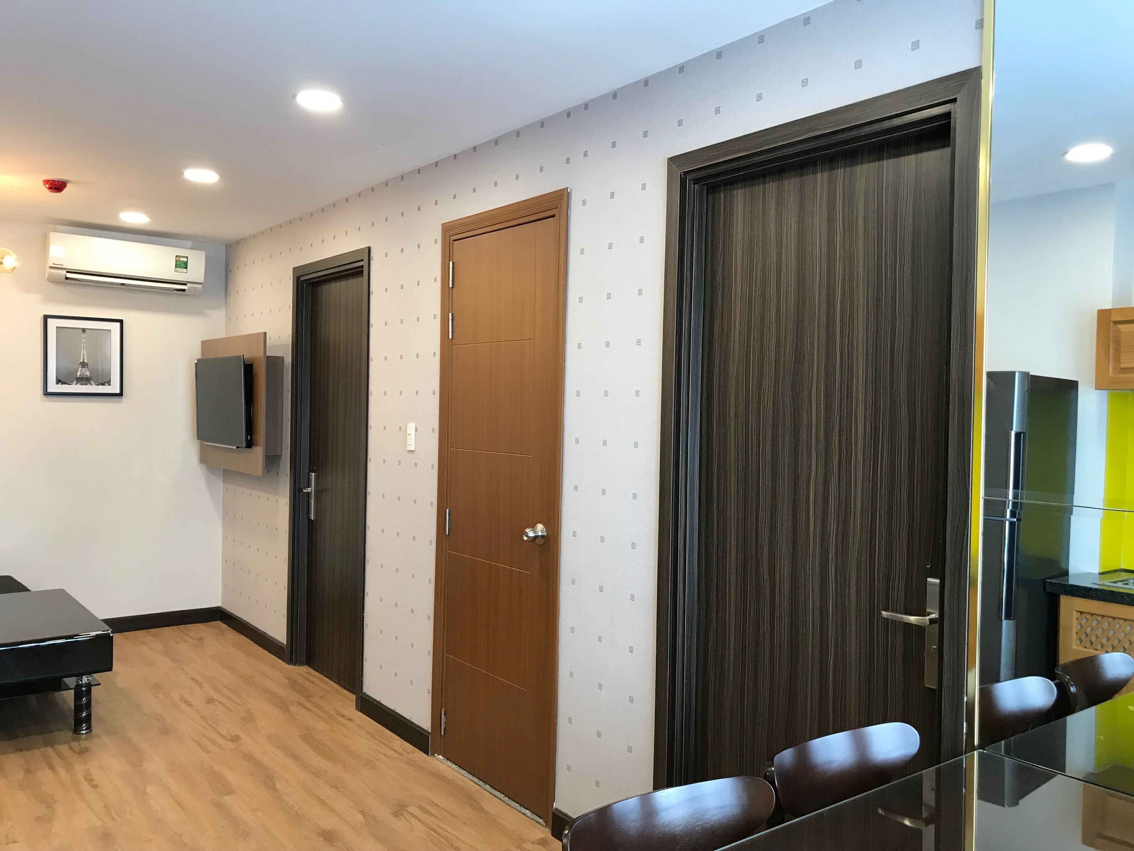 A modern hotel room with a wooden floor, a flat-screen TV mounted on the wall, a wooden door, and a glass partition with seating.