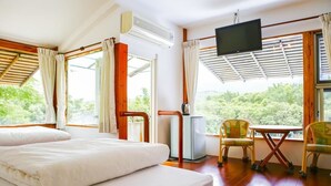 Habitación de hotel con cama, televisión, una mesita y sillas. Ventanas con cortinas y vistas al exterior con vegetación.