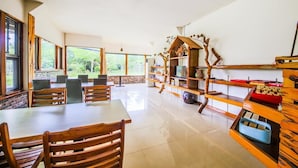 Un comedor amplio con mesas y sillas de madera, ventanas grandes que ofrecen vistas a la vegetación, y un estante rústico de madera con diversos objetos.