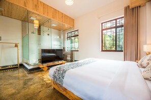 A modern bedroom with a large bed, a wooden ceiling, a glass shower enclosure, and a view of greenery through a window.