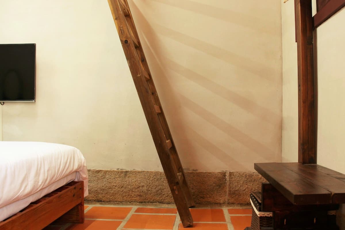A room with a bed, a wooden ladder leaning against the wall, a small table, and a television mounted on the wall.