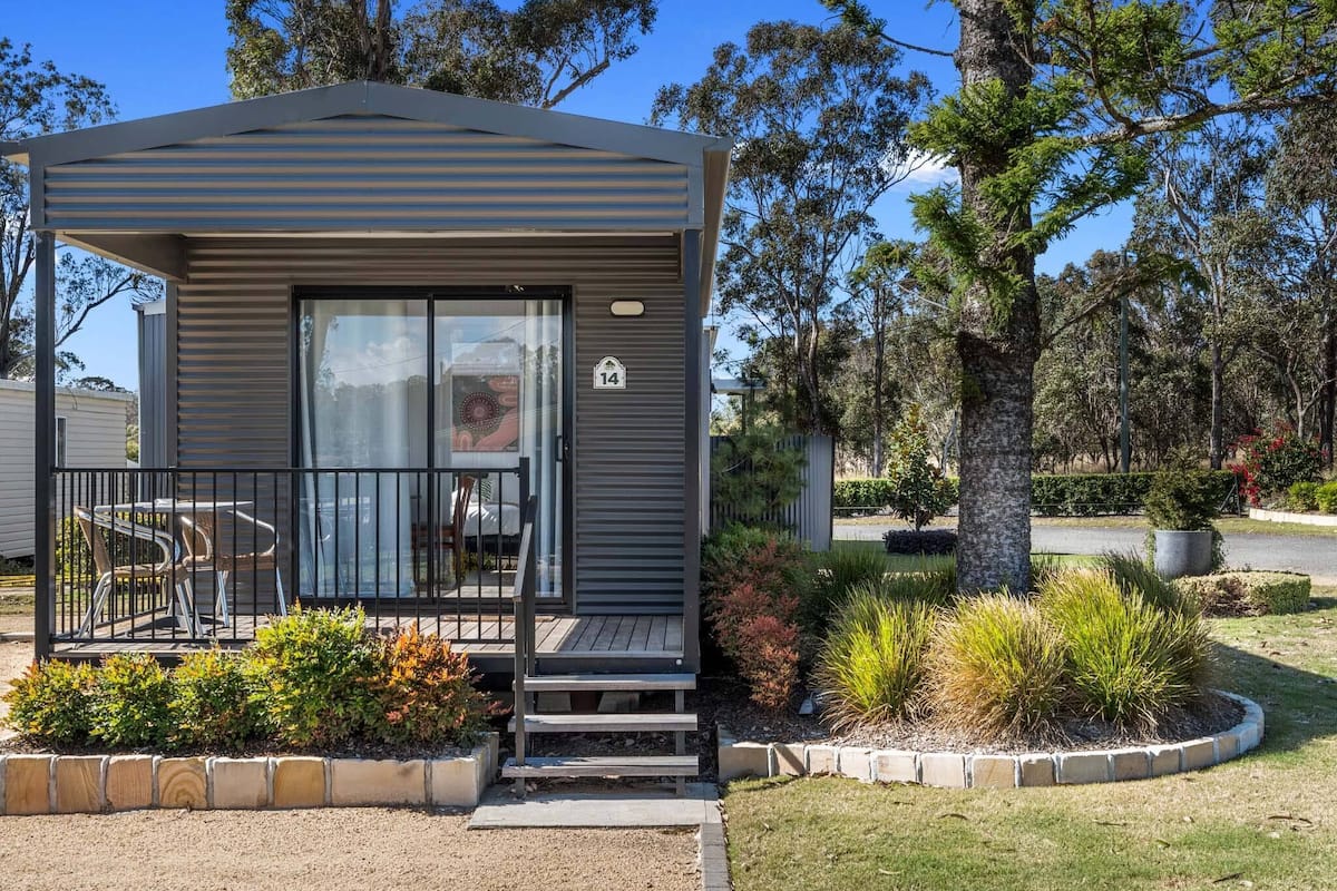 A small, single-story cabin with a balcony, surrounded by landscaped gardens and trees.