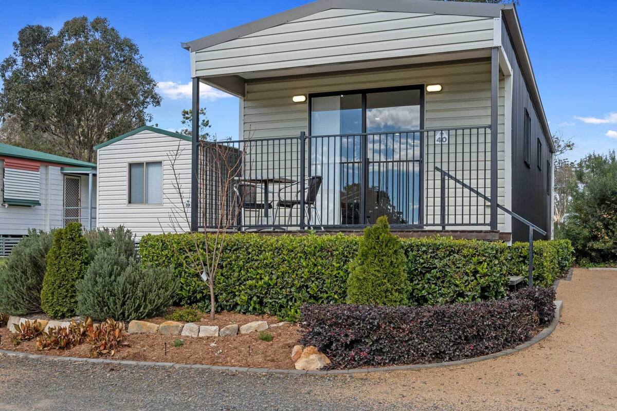 A modern two-story house with a balcony, a small garden area with various shrubs, and a paved driveway.