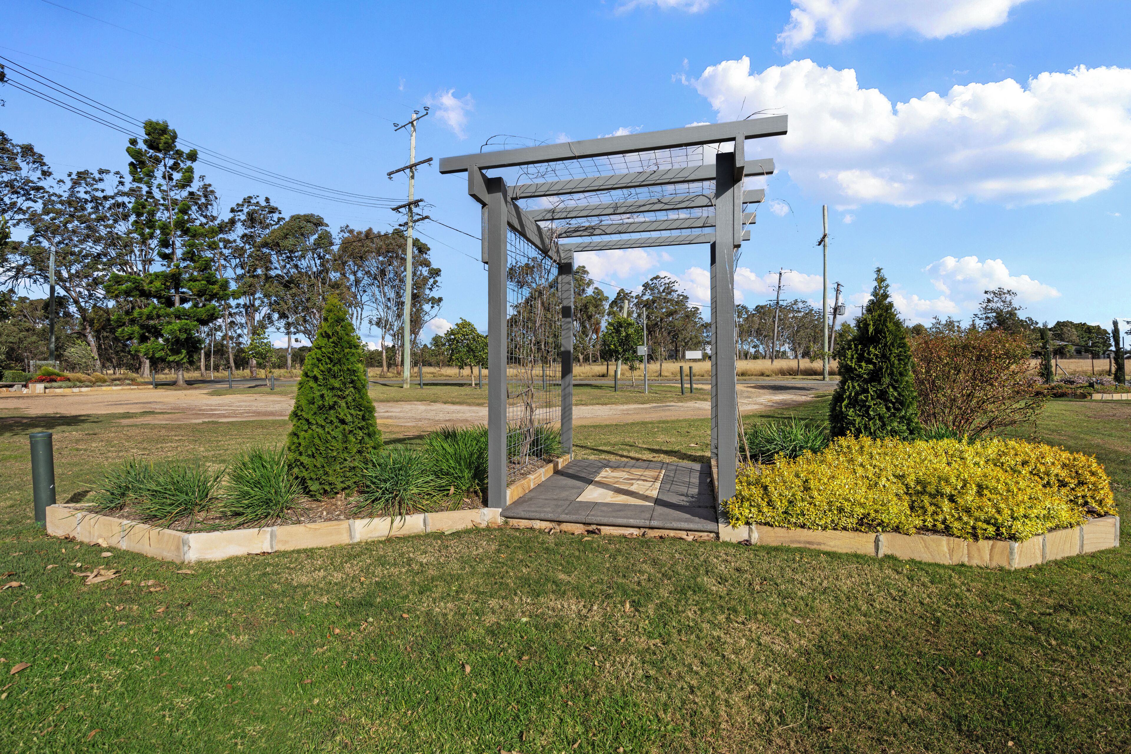 Una estructura de pérgola con armazón metálico y techo de malla de alambre, rodeada por un jardín ajardinado con diversas plantas y un camino empedrado que la conduce.