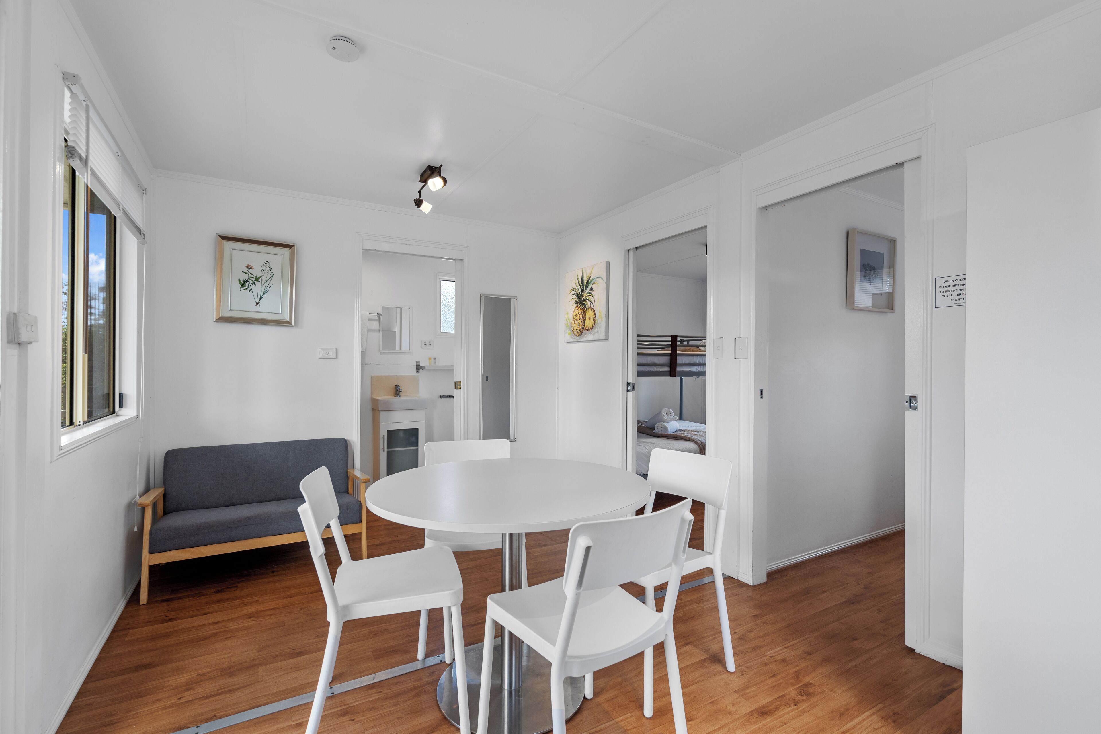 A modern, well-lit room with a round dining table, white chairs, a grey sofa, and wooden flooring.