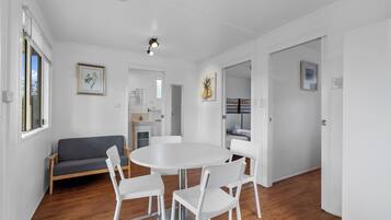A modern, well-lit room with a round dining table, white chairs, a grey sofa, and wooden flooring.