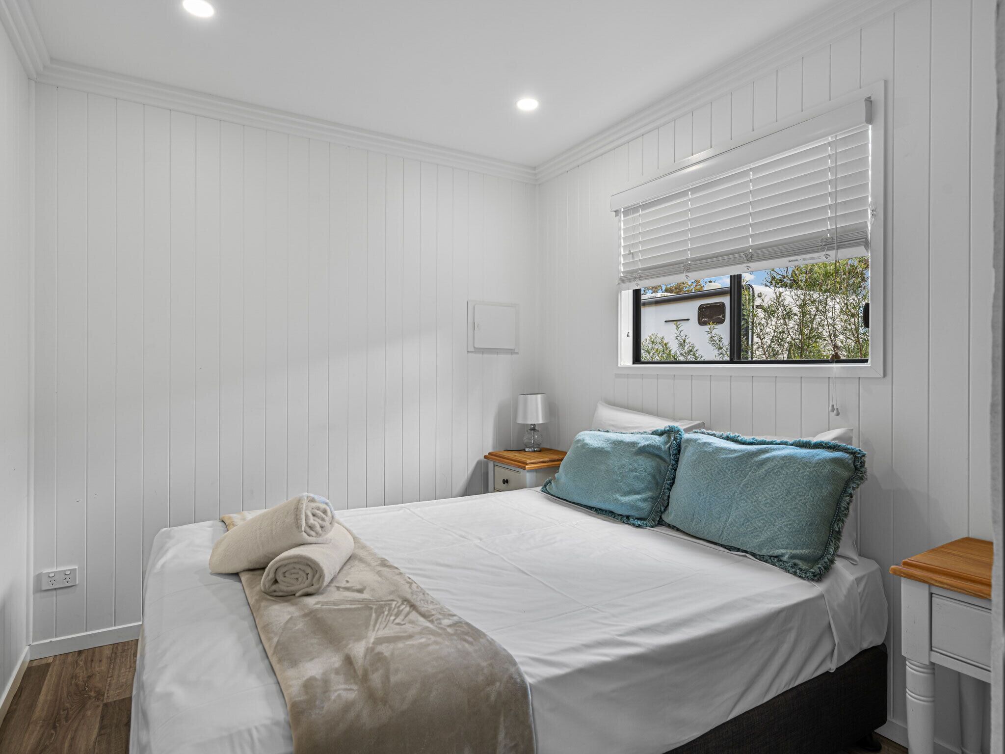 A bedroom with a bed, a nightstand, a window with blinds, and a view of the outside.
