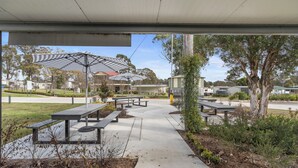 An outdoor patio area with picnic tables and umbrellas, surrounded by greenery and a pathway leading to a building. - Crows Nest Tourist Park (Crows Nest)