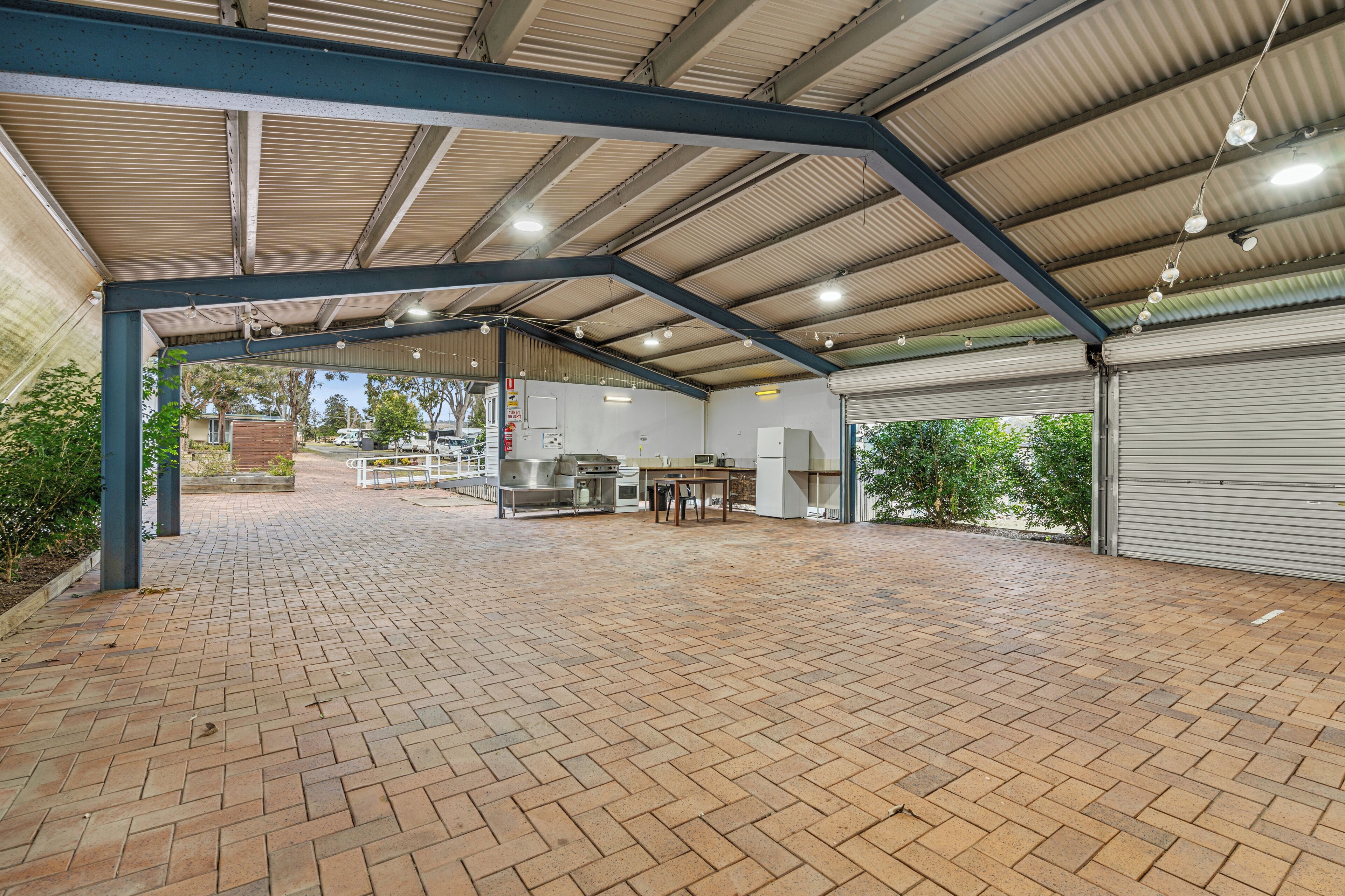 An open-air covered patio with a brick floor, metal beams, and a grill area.
