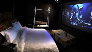 Une pièce faiblement éclairée, avec un lit, une petite table et un écran de projection sur lequel est diffusée une scène.