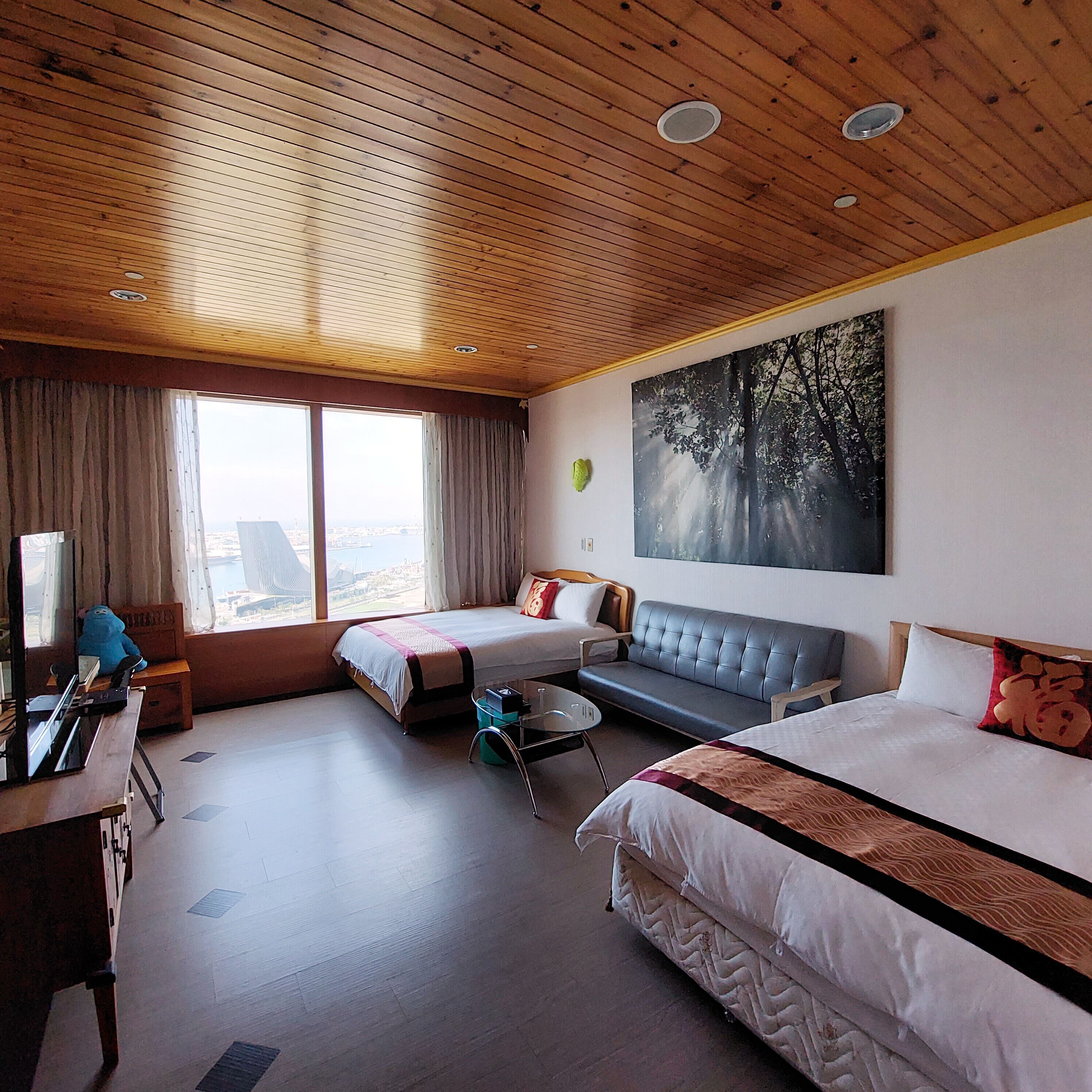 A hotel room with two beds, a sofa, a desk, and a large window with a view.
