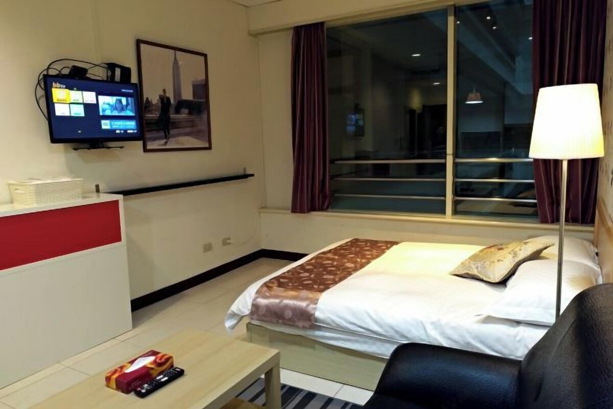 Una habitación de hotel con cama, escritorio, televisor y ventana con cortinas.