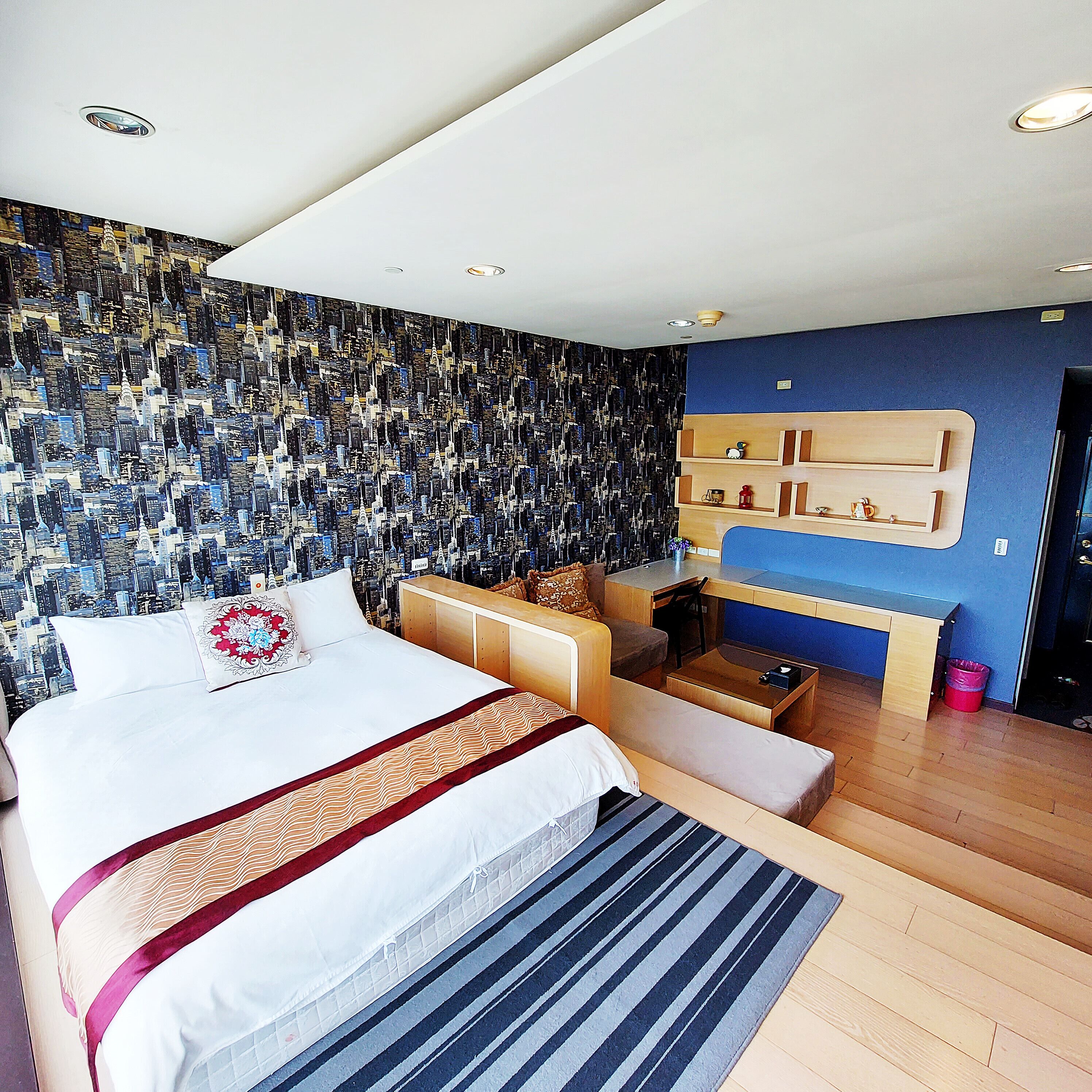 Un dormitorio moderno con papel tapiz con un paisaje urbano, una cama con colcha estampada, un escritorio y una silla.