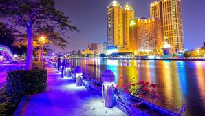 A nighttime view of a cityscape with two illuminated high-rise buildings by a calm river. - 85 Prague Homestay (Kaohsiung)