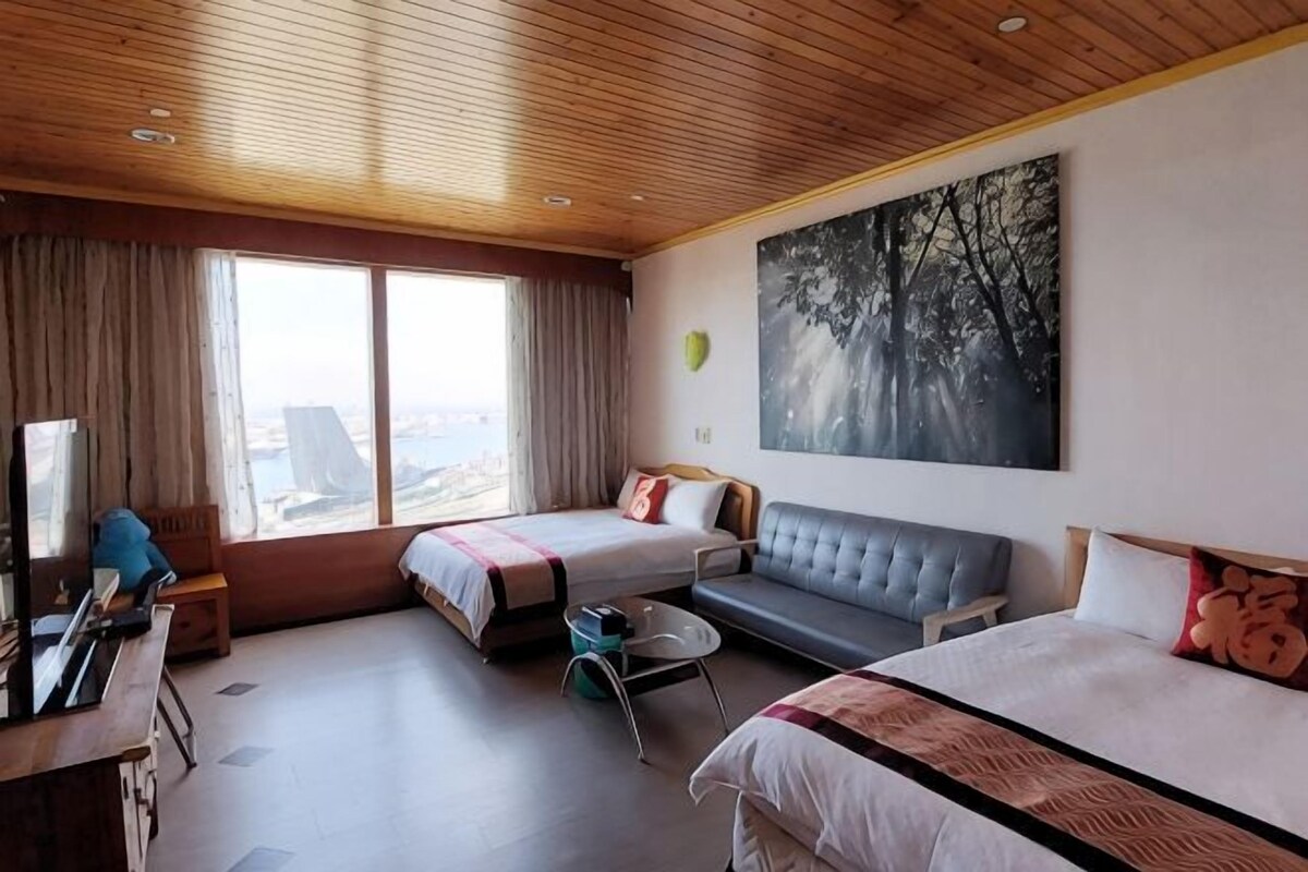 A hotel room with two beds, a sofa, a desk, and a large window with a view.