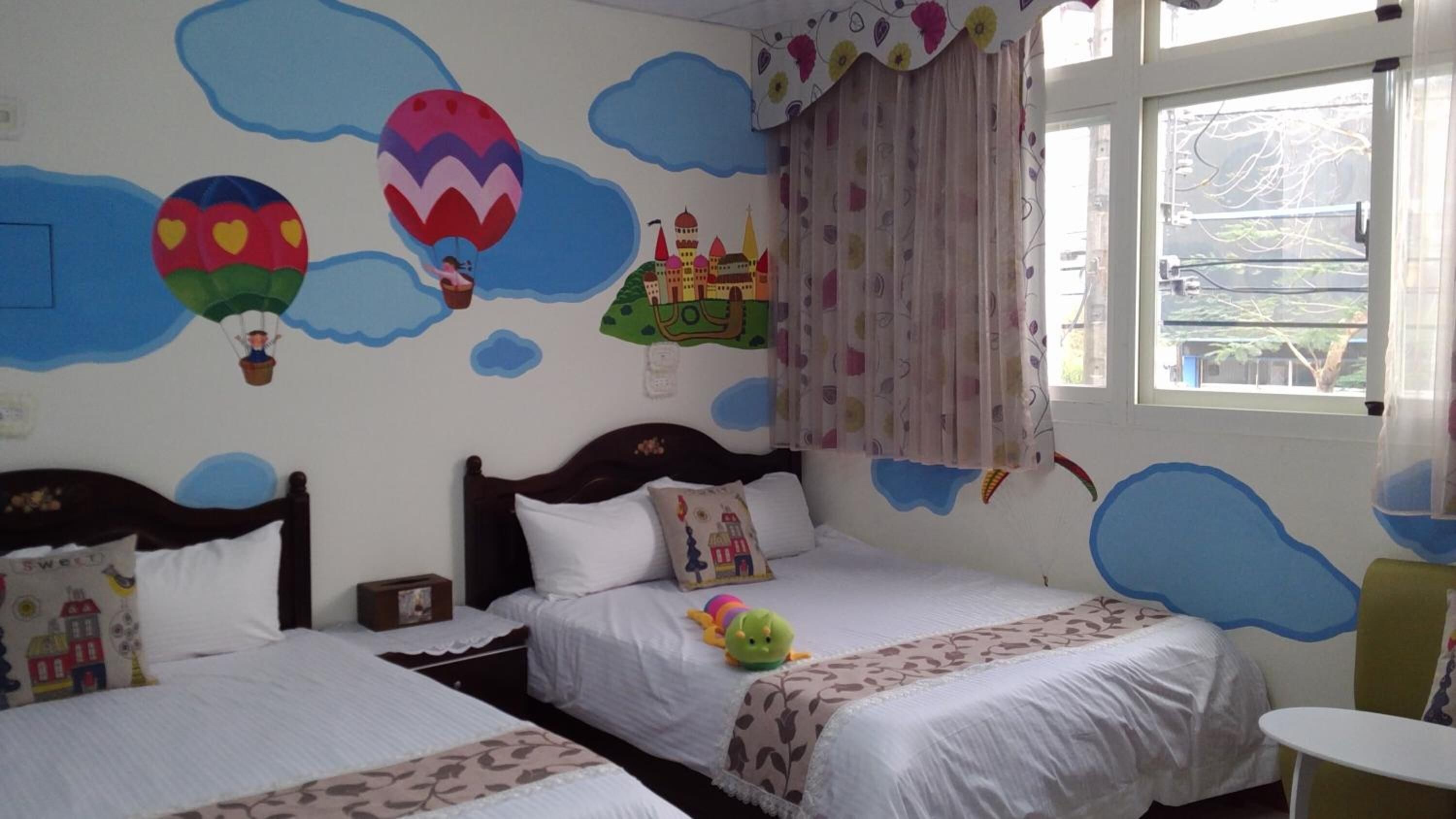 Una habitación con dos camas, una ventana con cortinas y una pared decorada con nubes y globos aerostáticos.