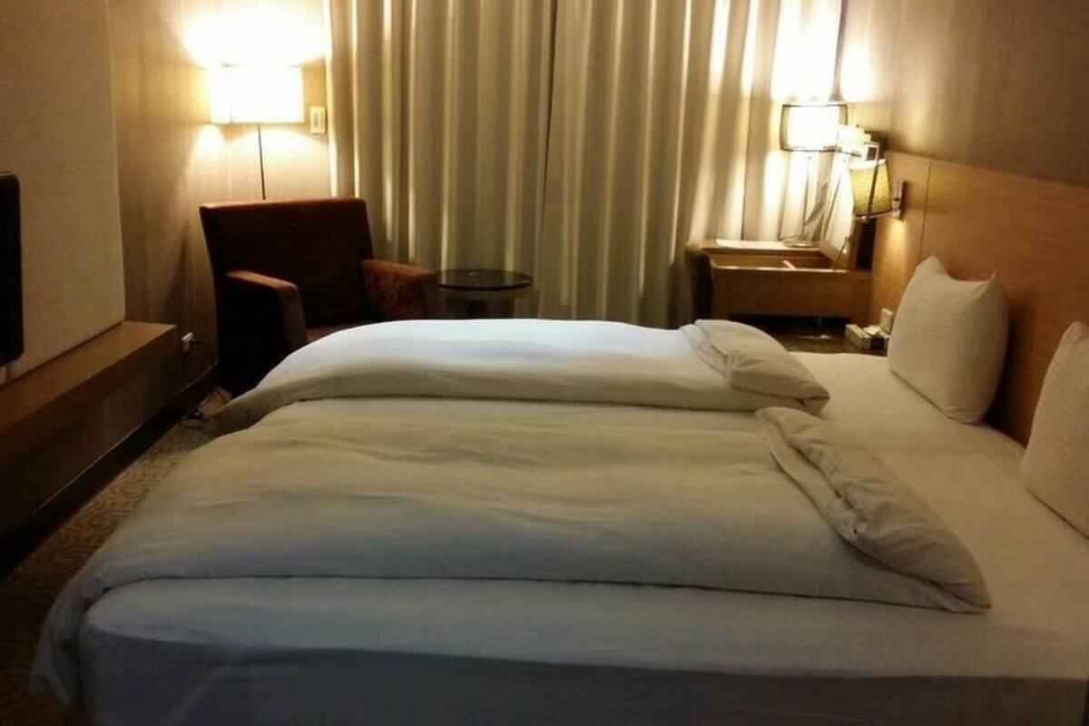 Quarto de hotel com uma cama grande, televisão, cadeira, mesa pequena e mesa de cabeceira com luminárias.
