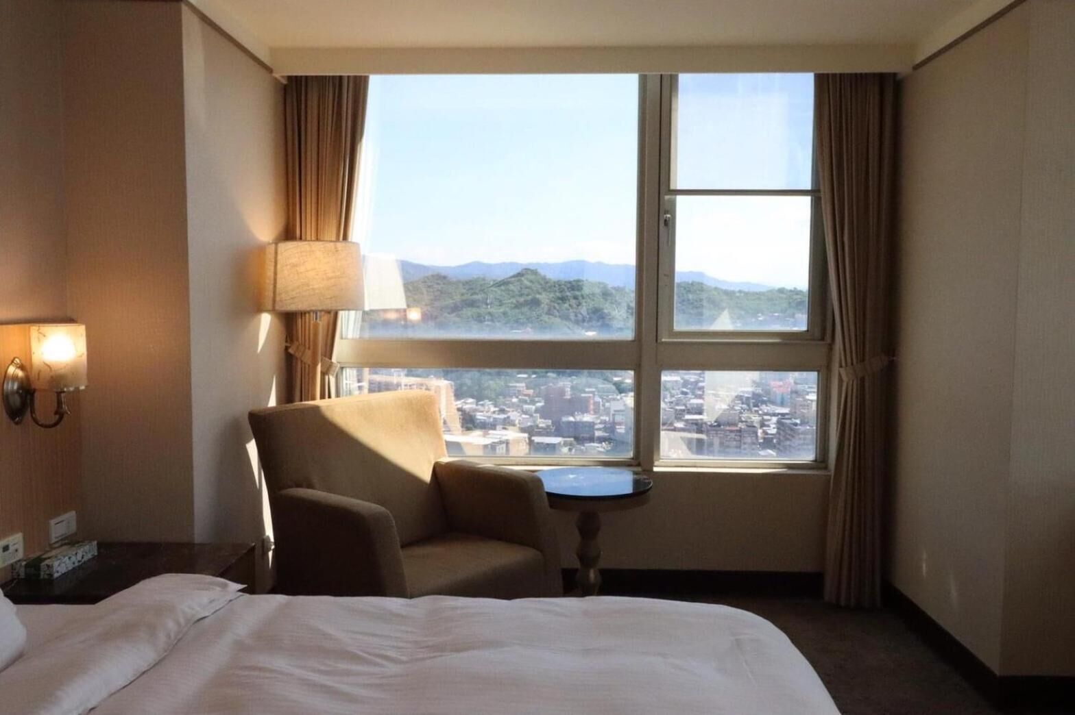 A hotel room with a bed, a chair, a small table, a lamp, and a large window with a city view.